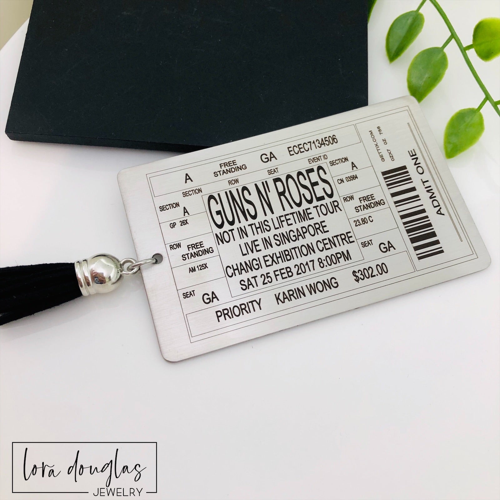 A stainless steel bookmark with laser engraved personalized text, depicting a concert ticket design, placed on a surface with a black bookmark and a green leaf in the background.
