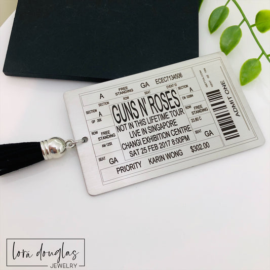 A stainless steel bookmark with laser engraved personalized text, depicting a concert ticket design, placed on a surface with a black bookmark and a green leaf in the background.