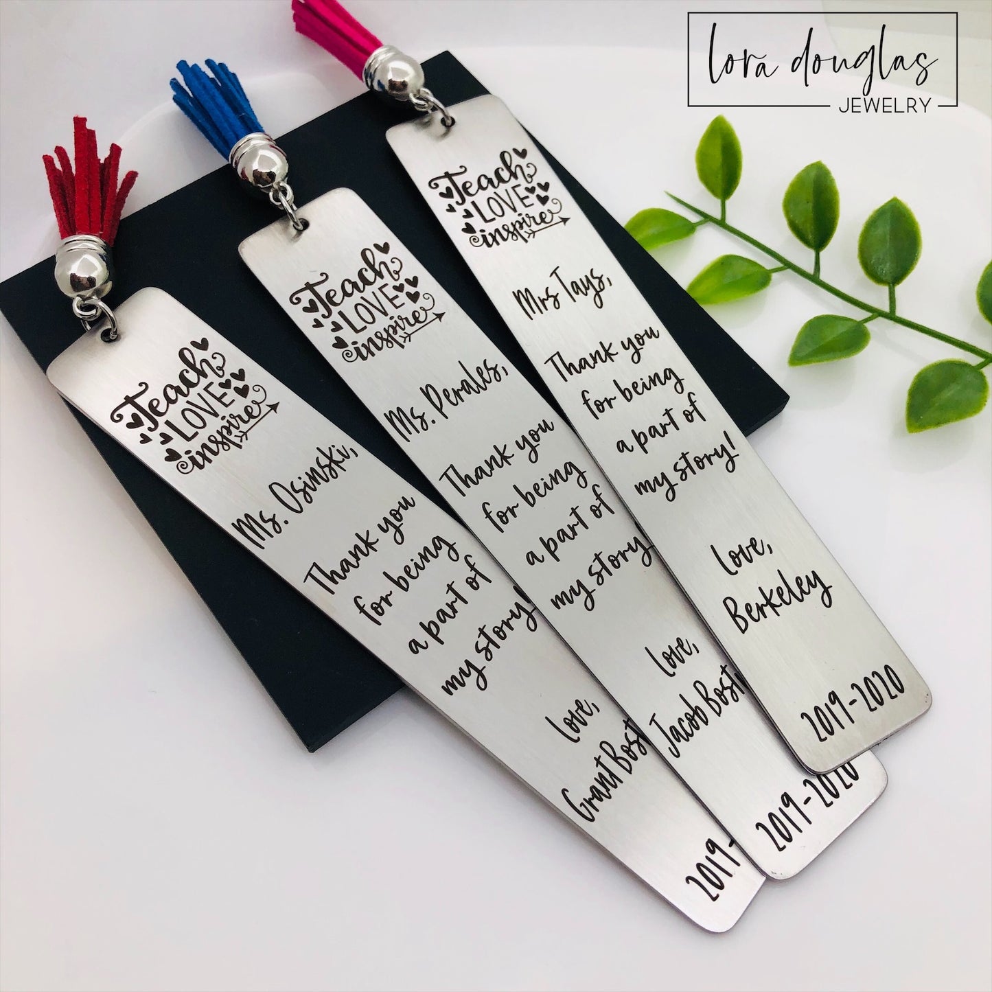 A set of stainless steel bookmarks with the phrases 'Teach Love Inspire' and 'Thank you for being my teacher' engraved on them, presented in a navy blue velvet pouch with tassels.