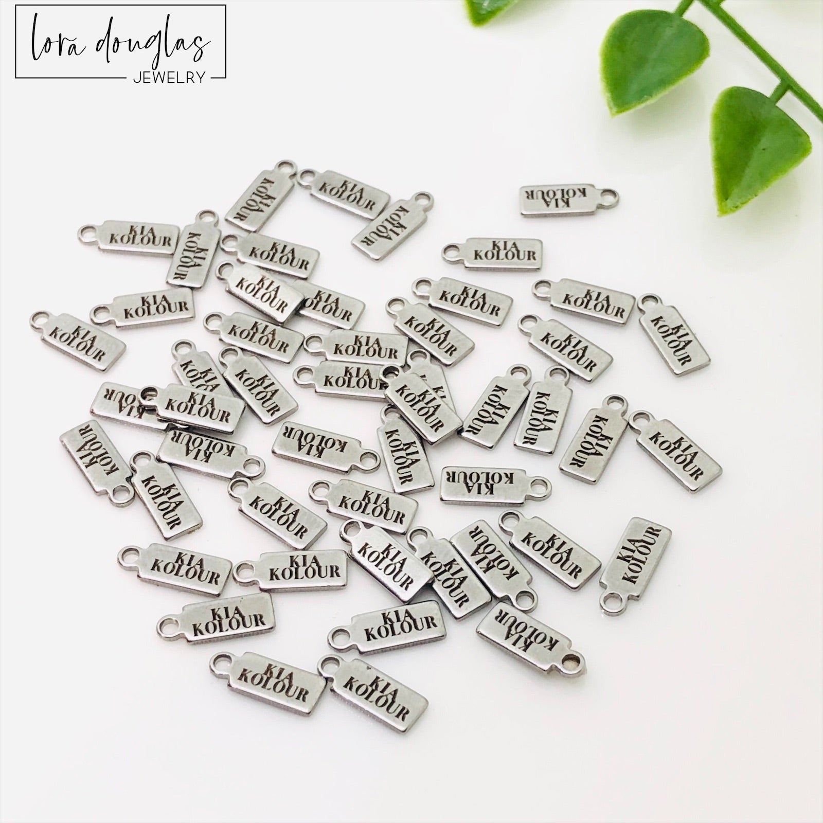 A collection of stainless steel jewelry tags with laser engraved logos and text on a white surface.