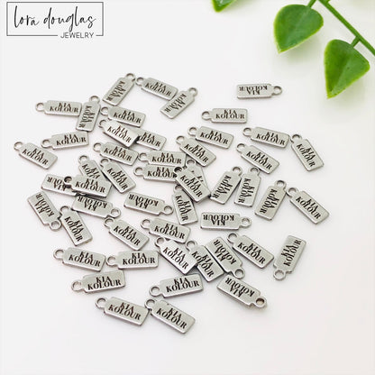 A collection of stainless steel jewelry tags with laser engraved logos and text on a white surface.