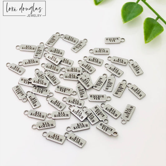 A collection of stainless steel jewelry tags with laser engraved logos and text on a white surface.