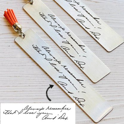 Two stainless steel bookmarks with engraved handwriting, with tassels, displayed on a wooden surface. The text appears to be a memorial message.