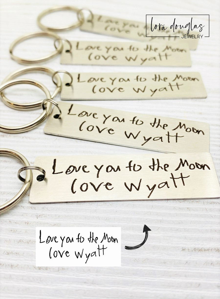 A stainless steel keychain with engraved handwriting samples. The keychain features a silver-colored plate with the text 'I love you dad' and 'Mom you're the best' alongside a standard key ring.