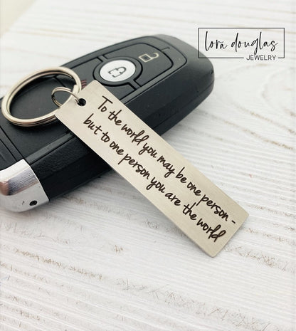 A stainless steel keychain with laser engraved text 'To The World You May Be One, But To One You Are The World' accompanied by a standard key ring, placed on a car key.