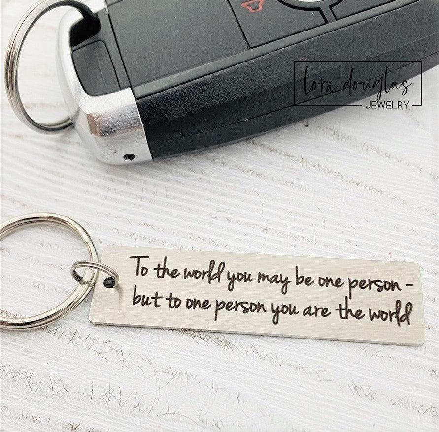 A stainless steel keychain with laser engraved text 'To The World You May Be One, But To One You Are The World' accompanied by a standard key ring, placed on a car key.