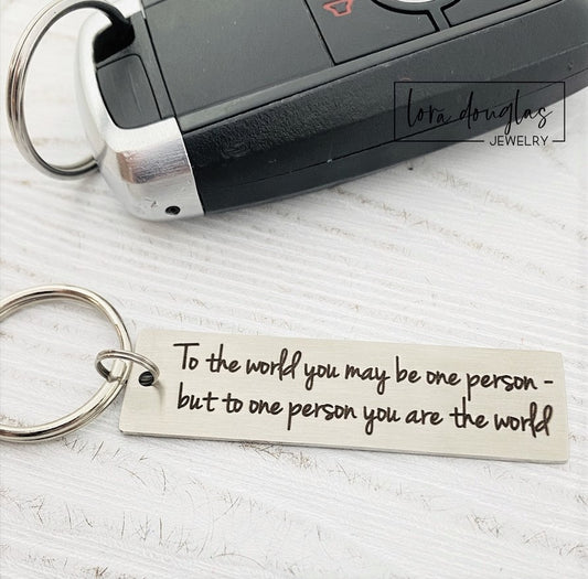 A stainless steel keychain with laser engraved text 'To The World You May Be One, But To One You Are The World' accompanied by a standard key ring, placed on a car key.