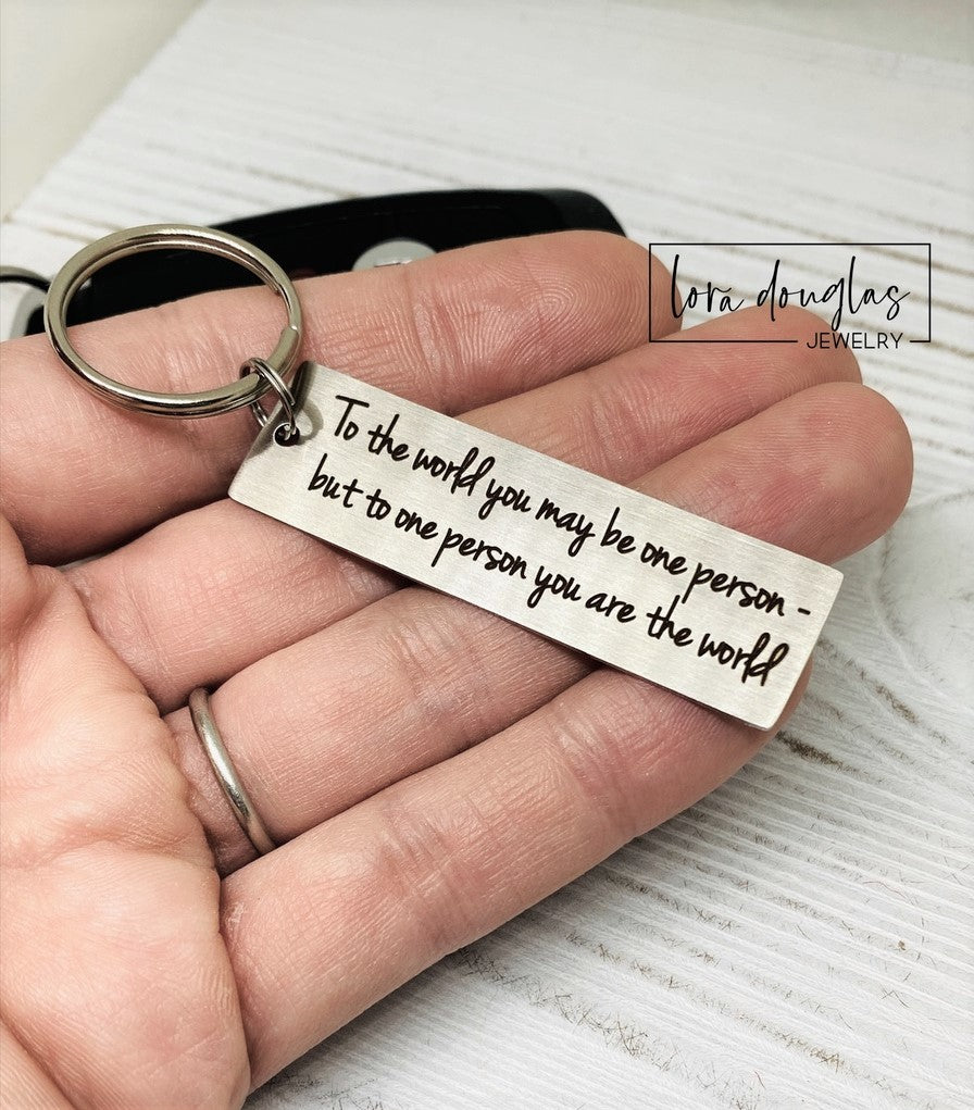 A stainless steel keychain with laser engraved text 'To The World You May Be One, But To One You Are The World' accompanied by a standard key ring, placed on a car key.