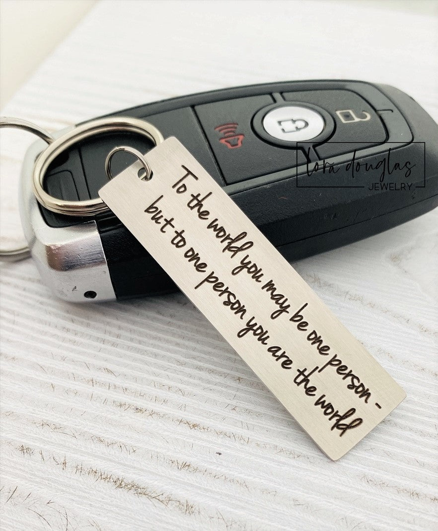A stainless steel keychain with laser engraved text 'To The World You May Be One, But To One You Are The World' accompanied by a standard key ring, placed on a car key.