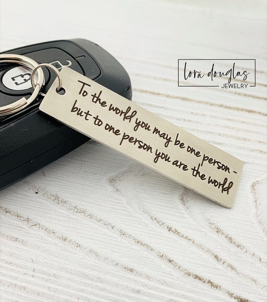 A stainless steel keychain with laser engraved text 'To The World You May Be One, But To One You Are The World' accompanied by a standard key ring, placed on a car key.