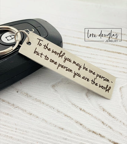 A stainless steel keychain with laser engraved text 'To The World You May Be One, But To One You Are The World' accompanied by a standard key ring, placed on a car key.