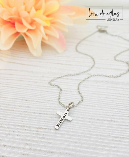 A silver stainless steel engraved cross necklace with a lobster clasp and a small cross charm, placed on a white surface with a few green leaves and a flower for decoration.
