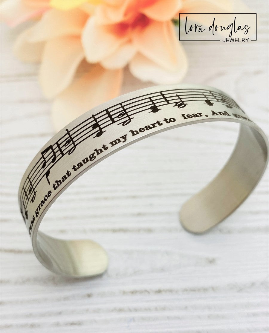 A stainless steel bracelet with sheet music engraved on it, displayed on a black surface with a green leafy element to the side.