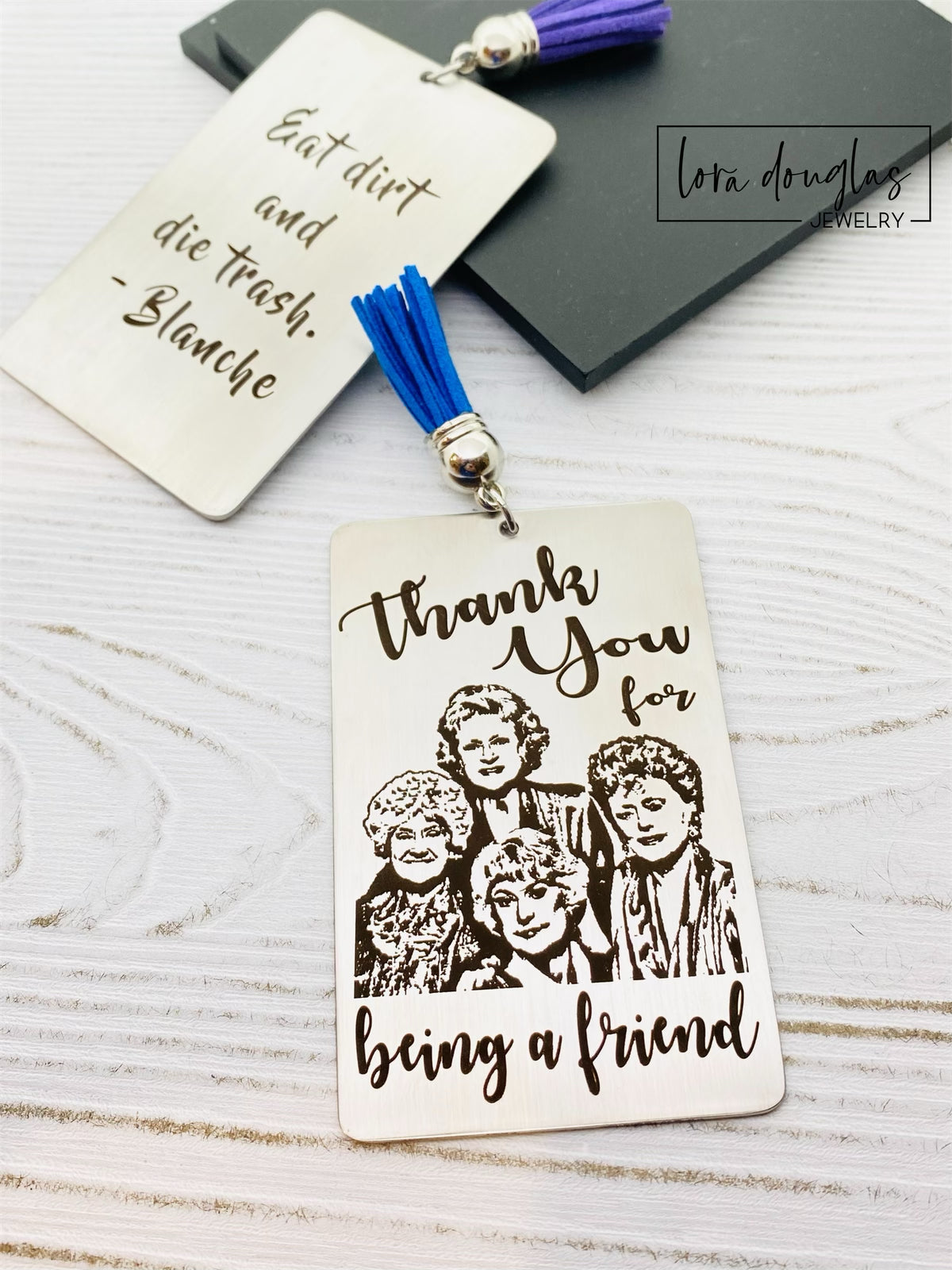 A stainless steel bookmark with engraved text and illustrations of the Golden Girls characters, with a navy blue velvet drawstring pouch and a gift tag.