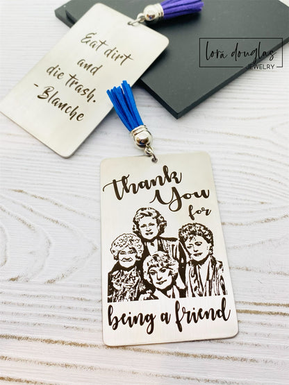 A stainless steel bookmark with engraved text and illustrations of the Golden Girls characters, with a navy blue velvet drawstring pouch and a gift tag.