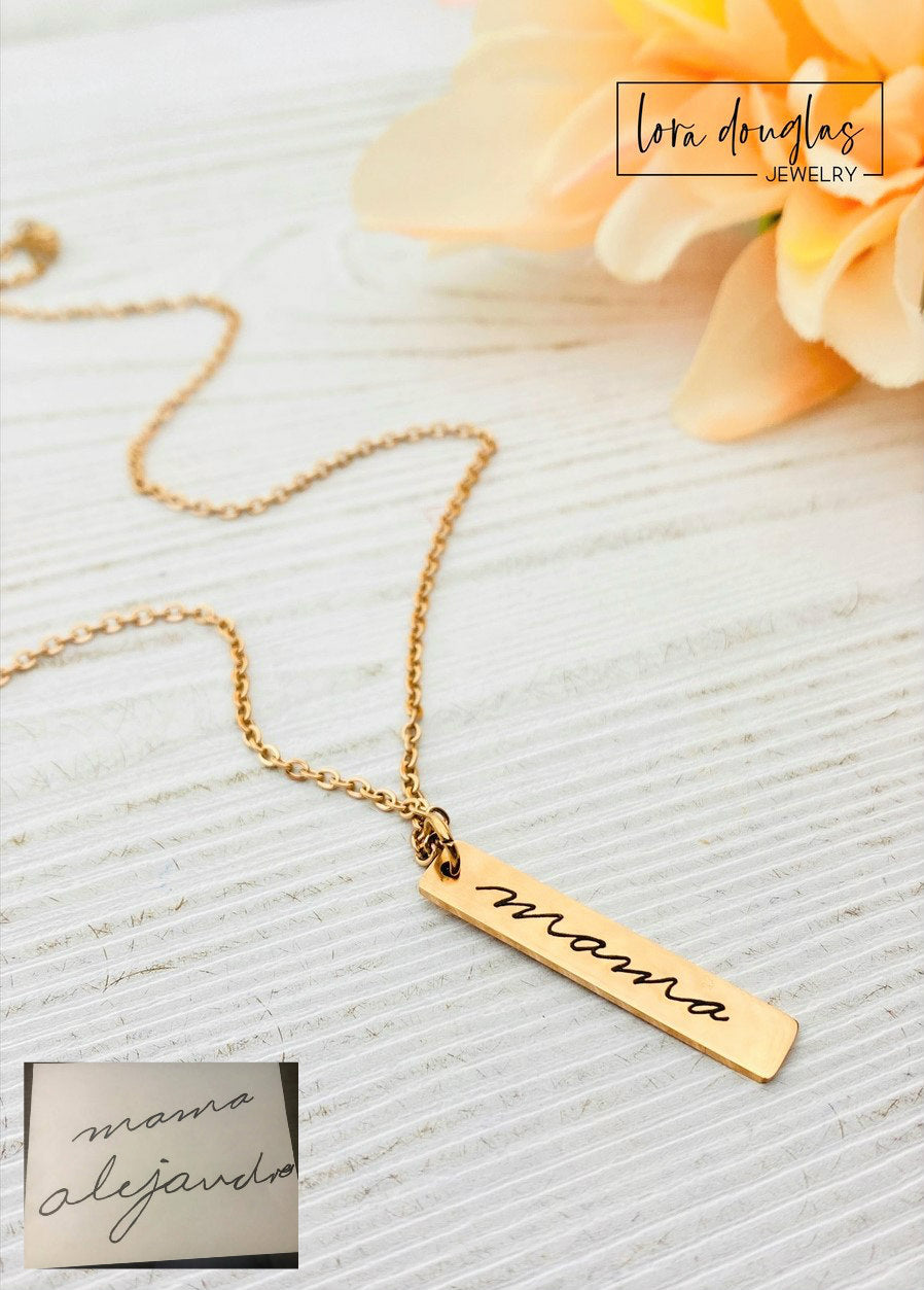 A gold stainless steel pendant with engraved handwriting, attached to an 18 inch chain with a lobster clasp, displayed against a white background with a blurred background hinting at a floral arrangement.