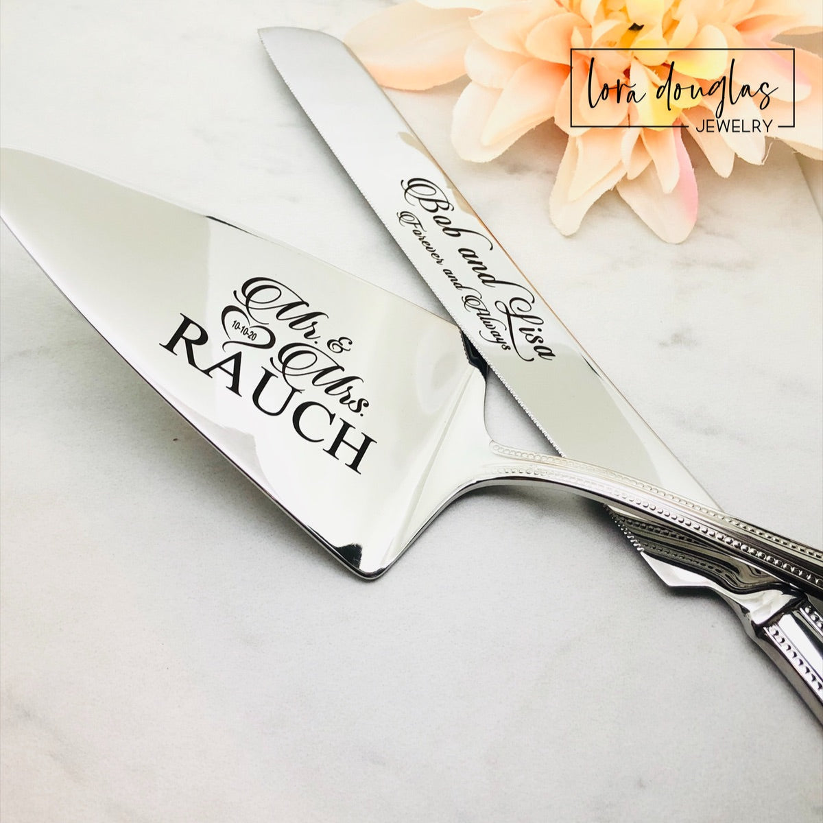 A set of two stainless steel cake serving tools, a knife and a server, with laser engraved personalization on the blade surfaces, placed on a marble surface with a floral decoration in the background.