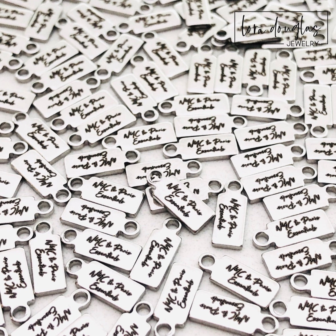 A collection of stainless steel jewelry tags with laser engraved logos and text on a white surface.