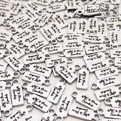A collection of stainless steel jewelry tags with laser engraved logos and text on a white surface.