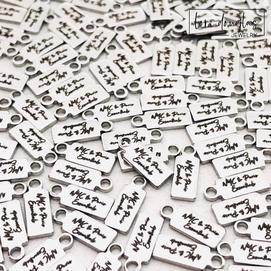 A collection of stainless steel jewelry tags with laser engraved logos and text on a white surface.