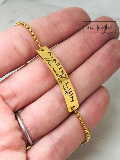 A gold bracelet with a bar pendant engraved with the handwritten message 