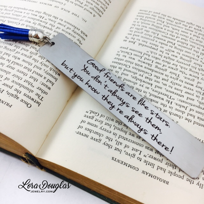 A stainless steel bookmark with an engraved message 'Good friends are like stars, you don't always see them but you know they're there.' placed on a book page, accompanied by a navy blue velvet drawstring pouch with a gift tag.