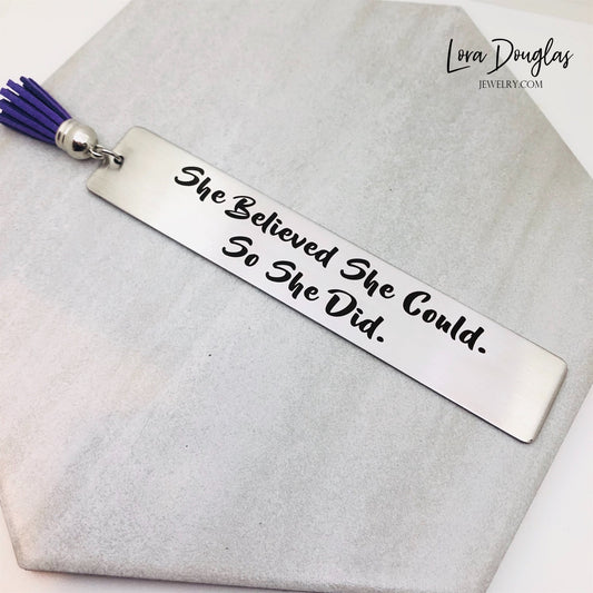 A stainless steel bookmark with the engraved text 'She Believed She Could So She Did' accompanied by a tassel.