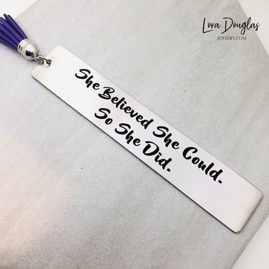 A stainless steel bookmark with the engraved text 'She Believed She Could So She Did' accompanied by a tassel.