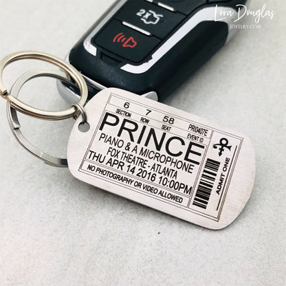 A stainless steel key chain with an engraved metal tag featuring text that resembles a Prince concert ticket. The key chain is shown with a car key.