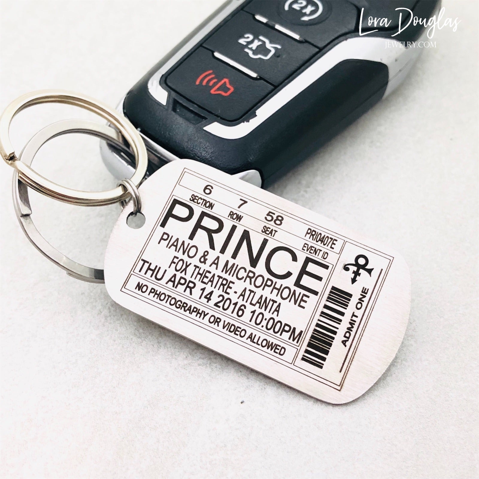 A stainless steel key chain with an engraved metal tag featuring text that resembles a Prince concert ticket. The key chain is shown with a car key.