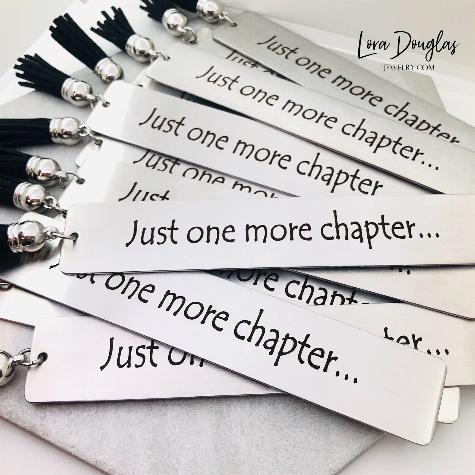 A stainless steel metal bookmark with the engraved text 'Just One More Chapter' and a tassel, placed on an open book.