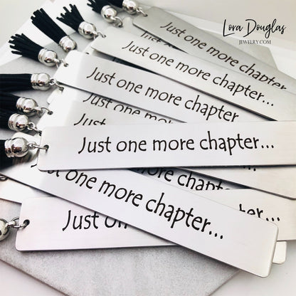 A stainless steel metal bookmark with the engraved text 'Just One More Chapter' and a tassel, placed on an open book.