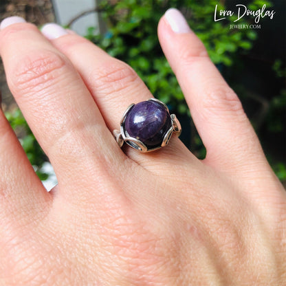 A hand displaying a sterling silver ring with a purple India ruby stone, held against a blurred background of greenery.