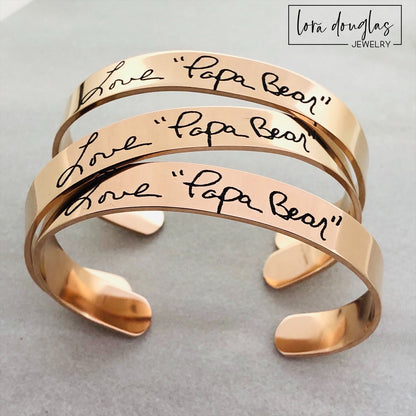 A stainless steel cuff bracelet with engraved handwriting on it, displayed on a light background.