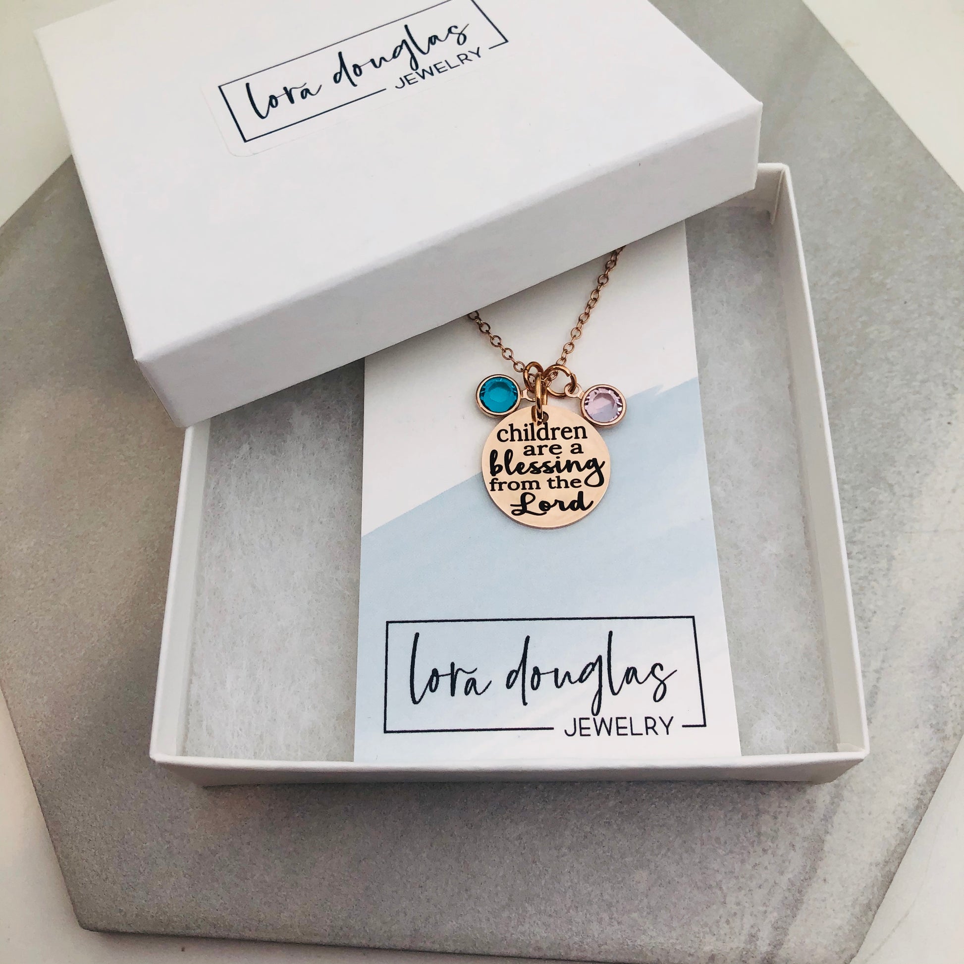 A rose gold stainless steel necklace with a round pendant that has the text 'CHILDREN ARE A BLESSING FROM THE LORD' engraved on it, along with a Swarovski Channel Plated Birthstone Charm.
