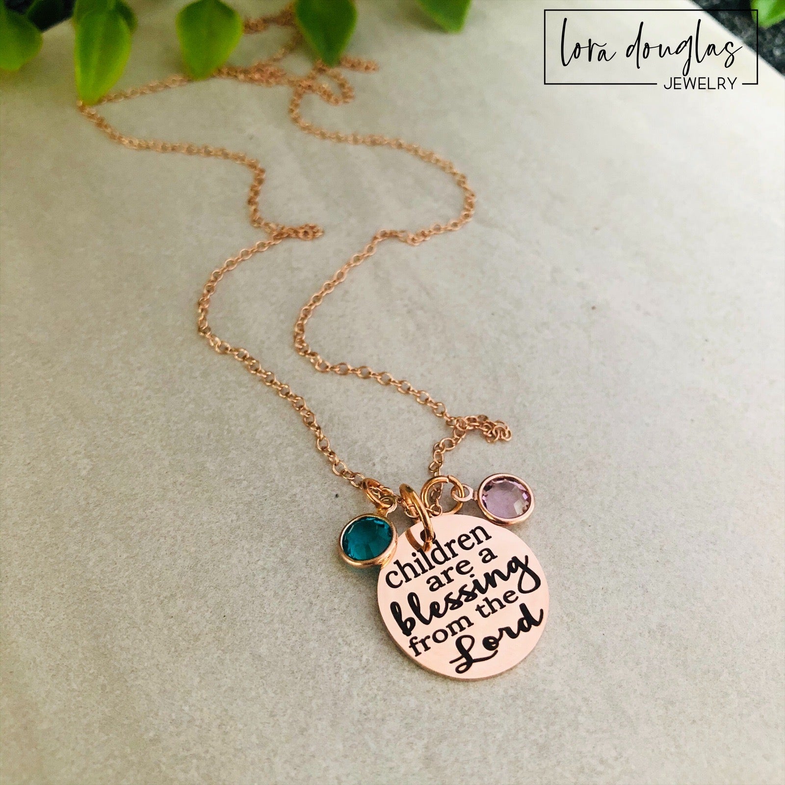 A rose gold stainless steel necklace with a round pendant that has the text 'CHILDREN ARE A BLESSING FROM THE LORD' engraved on it, along with a Swarovski Channel Plated Birthstone Charm.