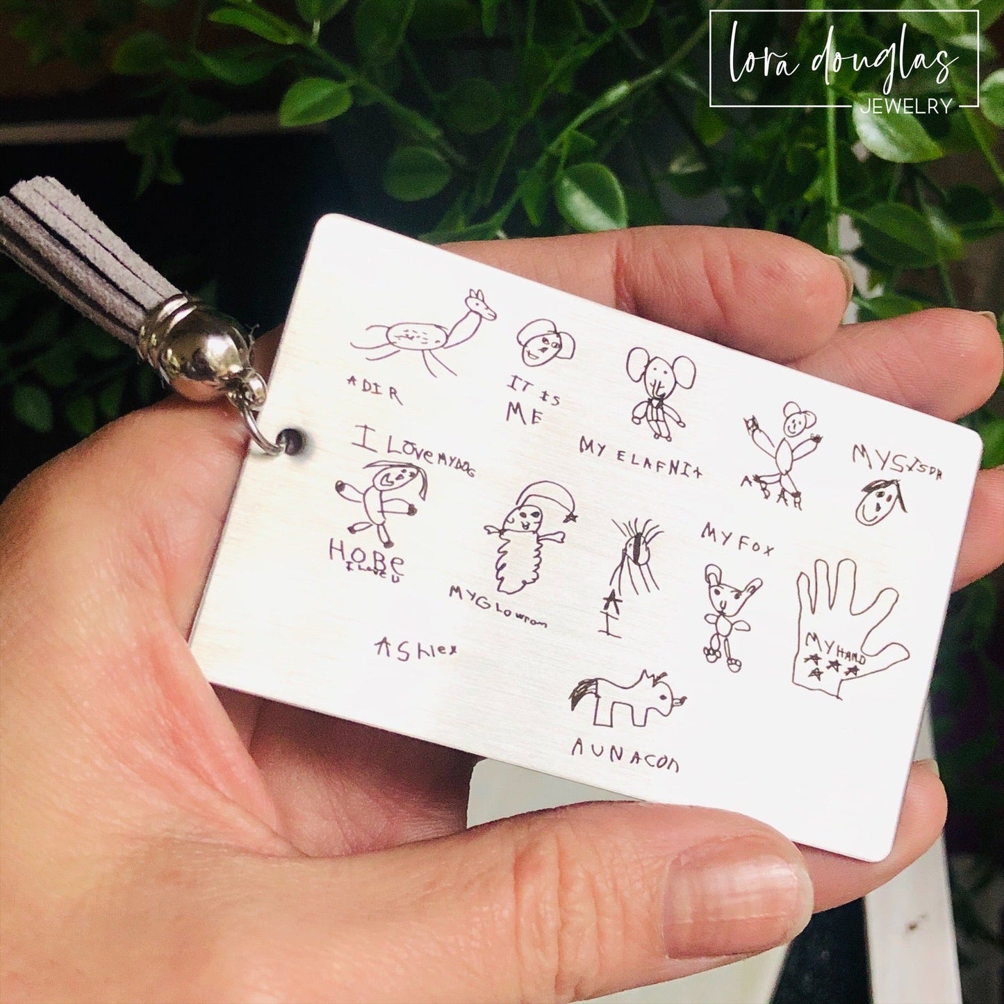 A custom engraved bookmark with a child's drawing engraved on the metal. The bookmark is silver with black engraving and has a navy blue Tassel.