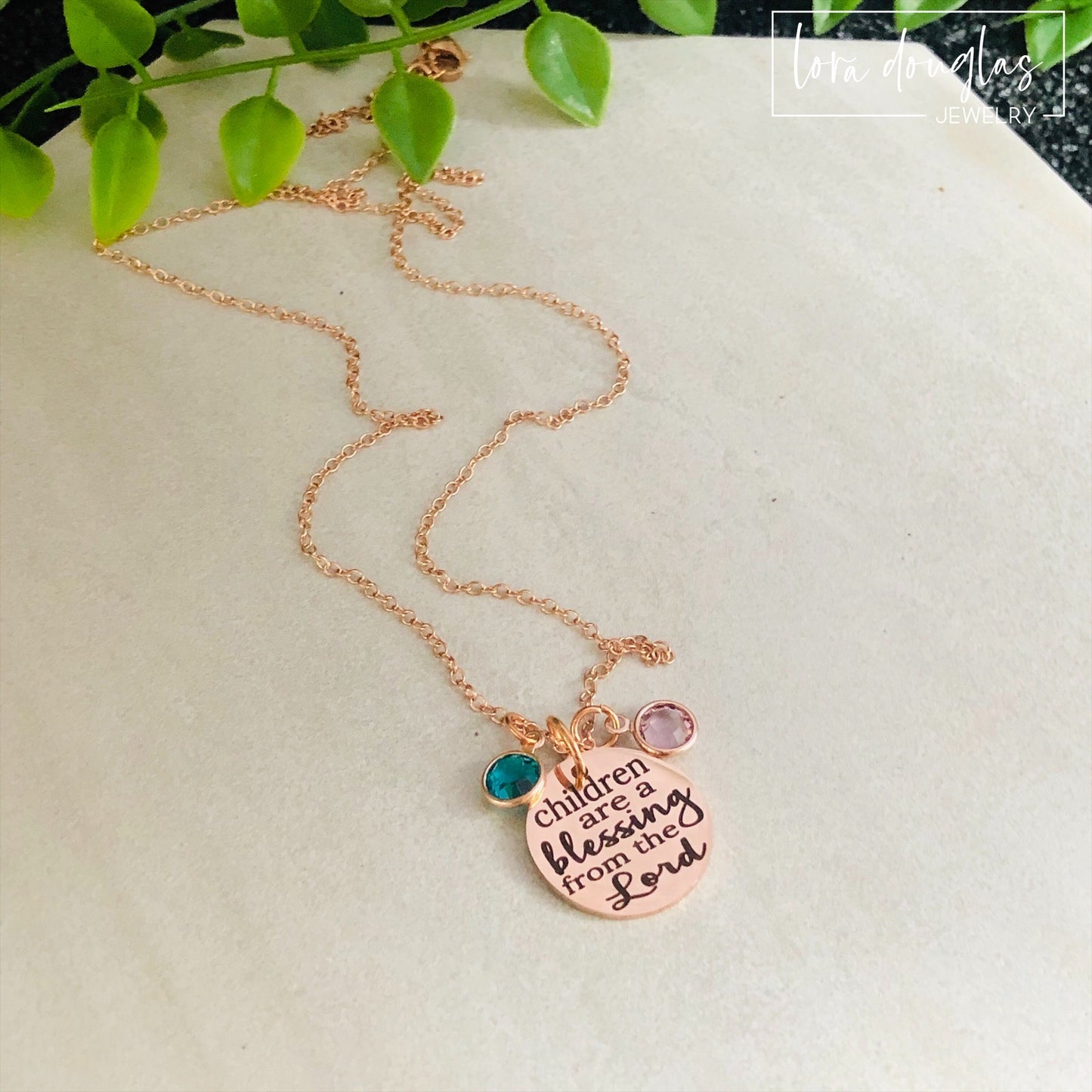 A rose gold stainless steel necklace with a round pendant that has the text 'CHILDREN ARE A BLESSING FROM THE LORD' engraved on it, along with a Swarovski Channel Plated Birthstone Charm.