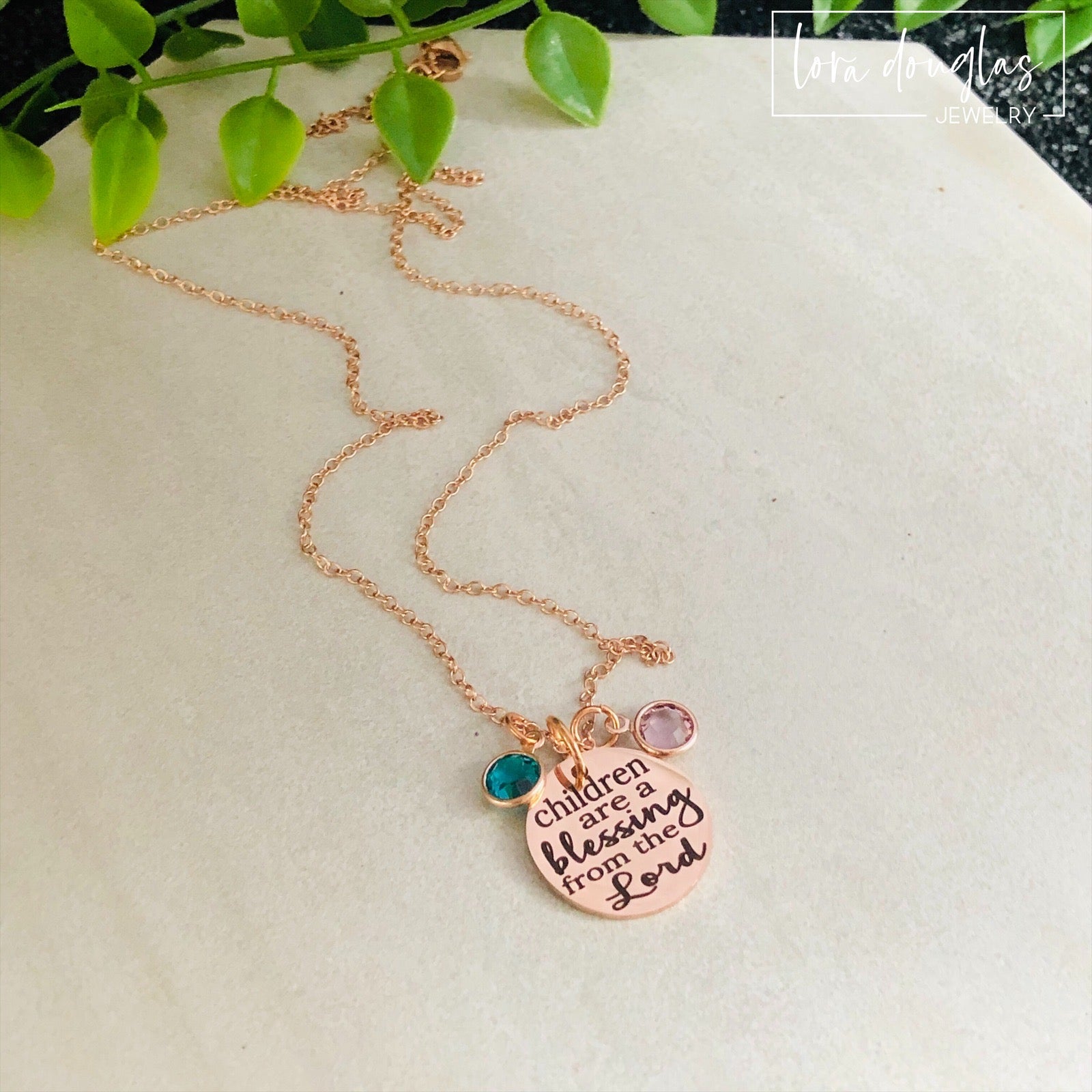 A rose gold stainless steel necklace with a round pendant that has the text 'CHILDREN ARE A BLESSING FROM THE LORD' engraved on it, along with a Swarovski Channel Plated Birthstone Charm.