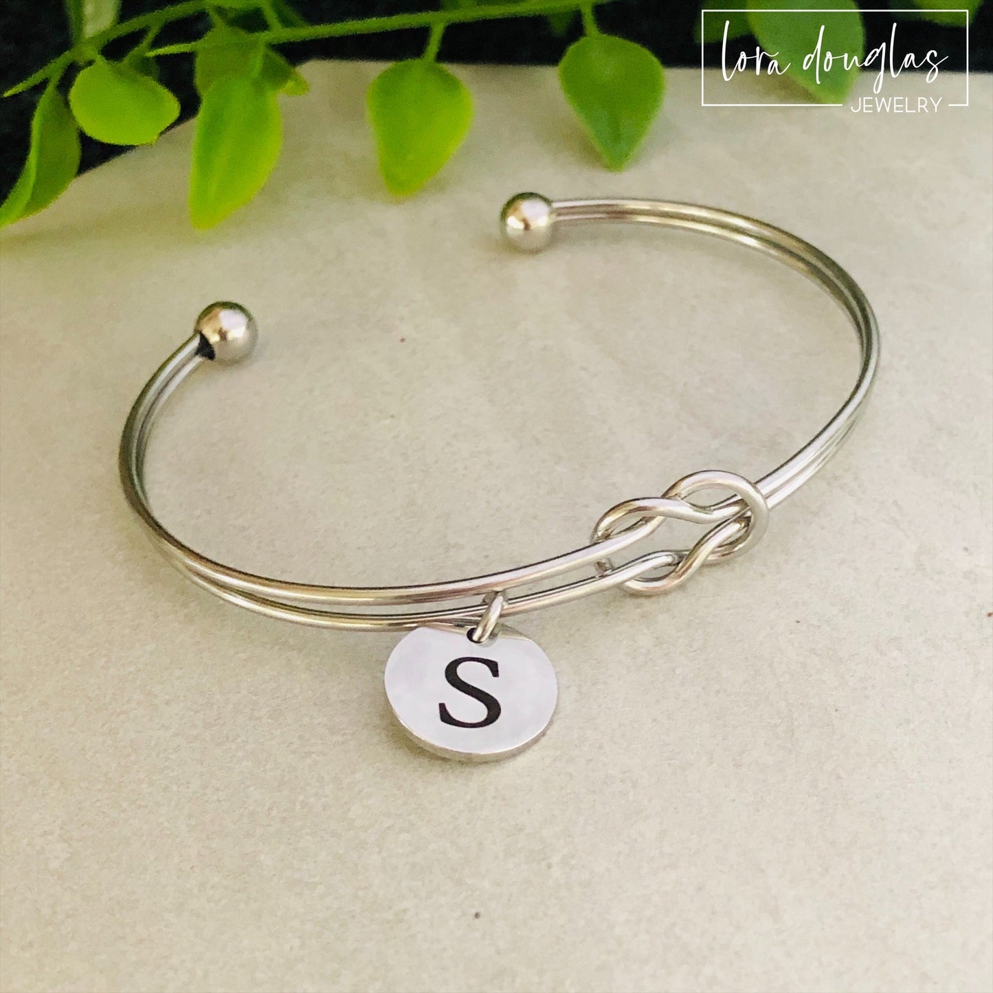 A silver stainless steel bracelet with a personalized initial disc charm engraved on a label, displayed on a flat surface with a green plant in the background.