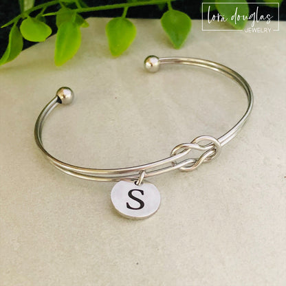 A silver stainless steel bracelet with a personalized initial disc charm engraved on a label, displayed on a flat surface with a green plant in the background.