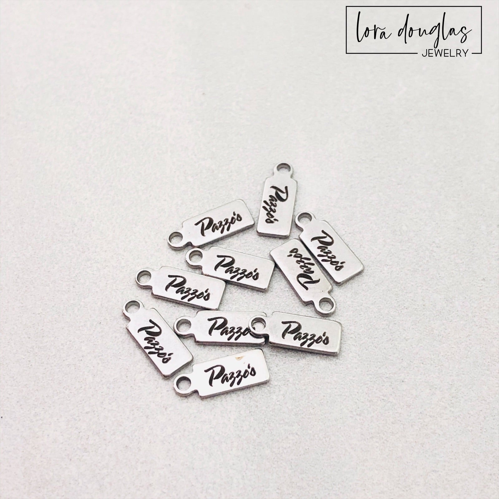 A collection of stainless steel jewelry tags with laser engraved logos and text on a white surface.