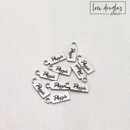 A collection of stainless steel jewelry tags with laser engraved logos and text on a white surface.