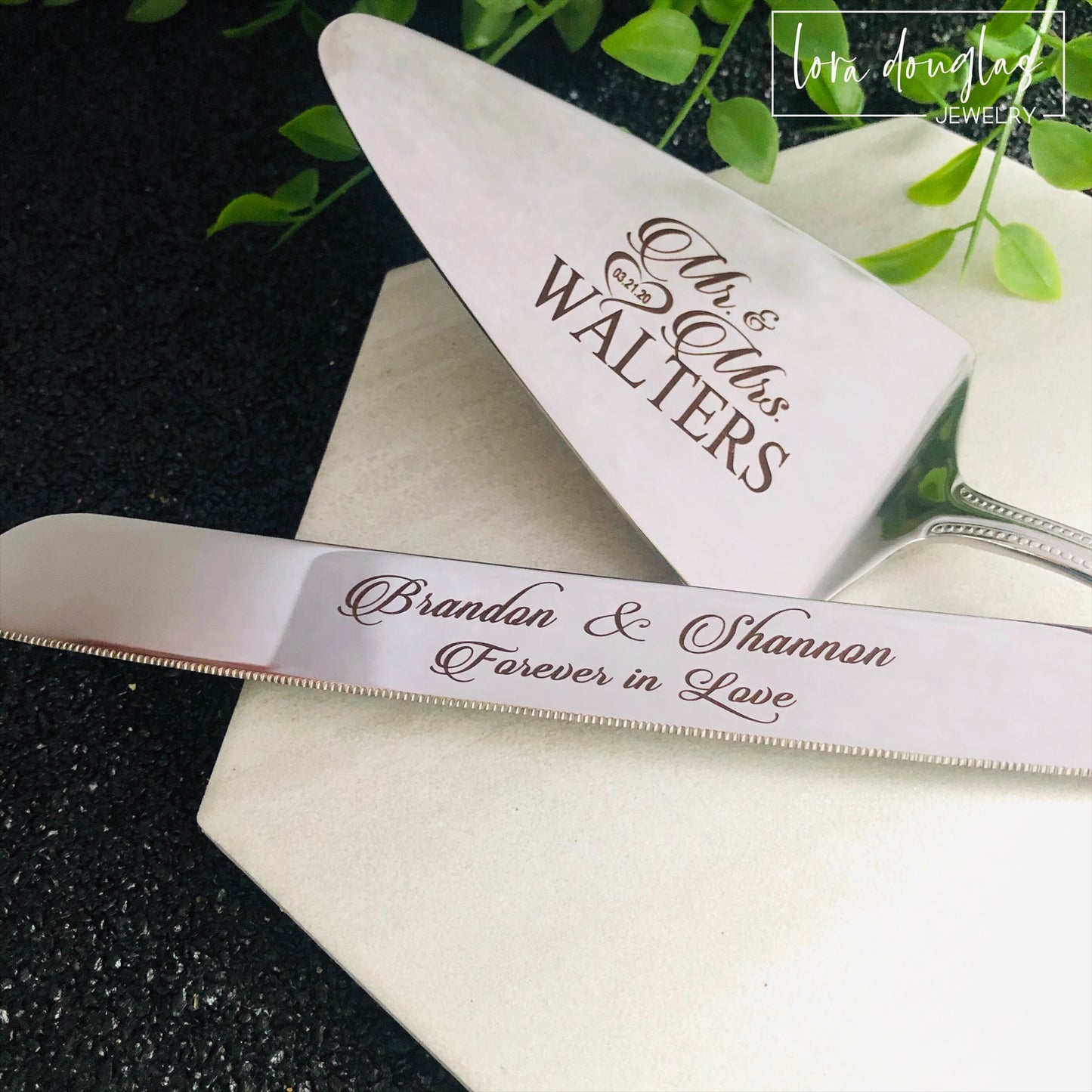 A set of two stainless steel cake serving tools, a knife and a server, with laser engraved personalization on the blade surfaces, placed on a marble surface with a floral decoration in the background.