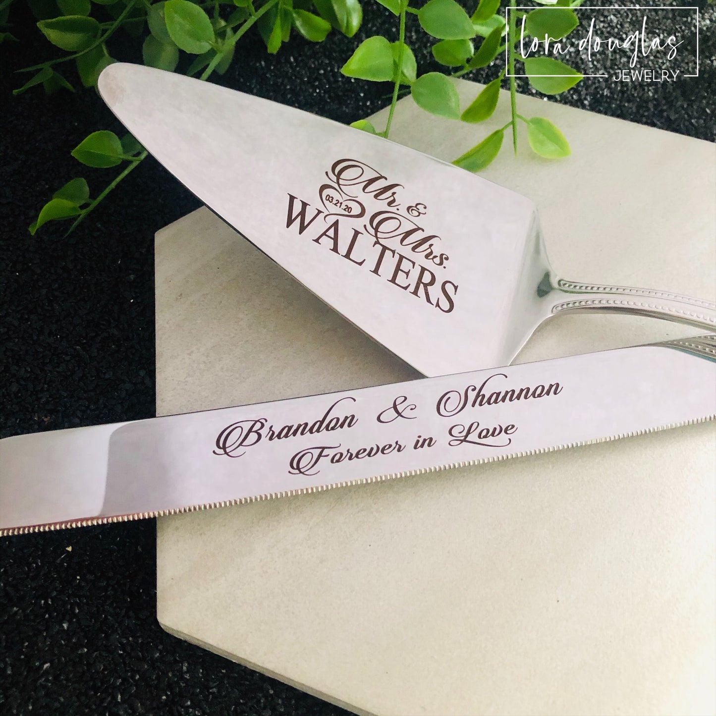 A set of two stainless steel cake serving tools, a knife and a server, with laser engraved personalization on the blade surfaces, placed on a marble surface with a floral decoration in the background.