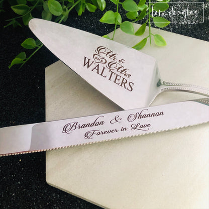 A set of two stainless steel cake serving tools, a knife and a server, with laser engraved personalization on the blade surfaces, placed on a marble surface with a floral decoration in the background.