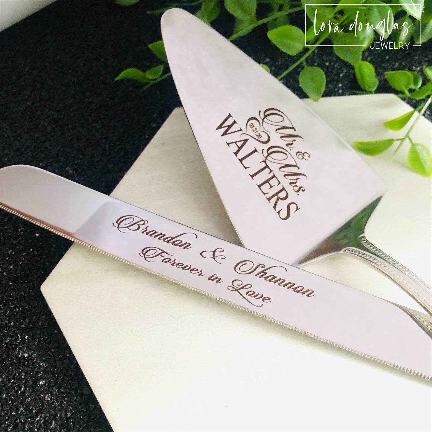 A set of two stainless steel cake serving tools, a knife and a server, with laser engraved personalization on the blade surfaces, placed on a marble surface with a floral decoration in the background.
