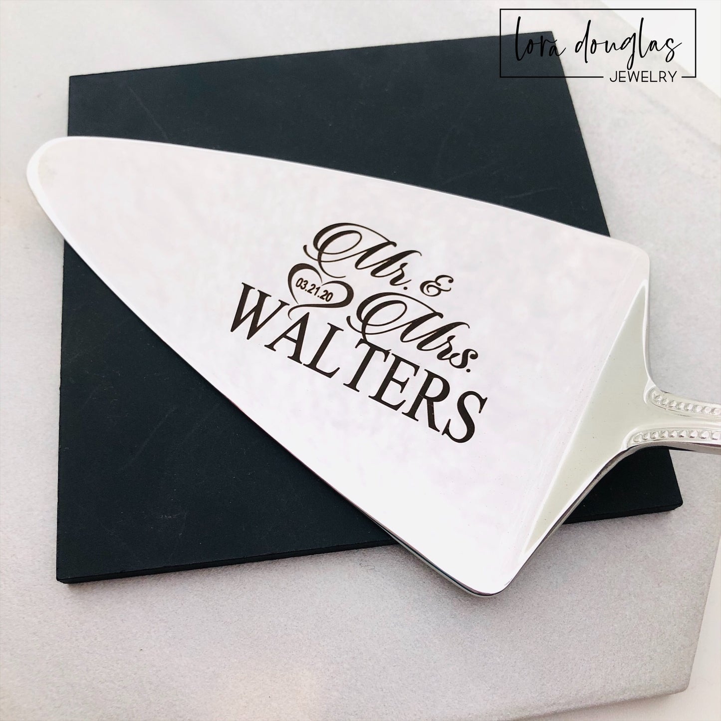 A set of two stainless steel cake serving tools, a knife and a server, with laser engraved personalization on the blade surfaces, placed on a marble surface with a floral decoration in the background.