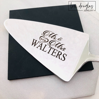 A set of two stainless steel cake serving tools, a knife and a server, with laser engraved personalization on the blade surfaces, placed on a marble surface with a floral decoration in the background.