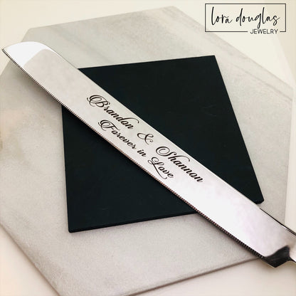 A set of two stainless steel cake serving tools, a knife and a server, with laser engraved personalization on the blade surfaces, placed on a marble surface with a floral decoration in the background.
